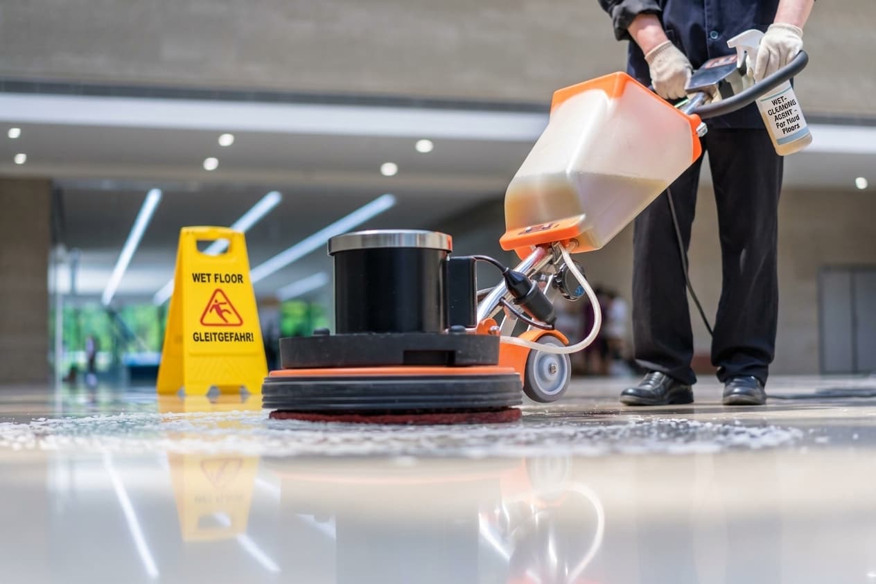 Reinigungskraft führt eine Einscheibenmaschine über den Steinboden einer Eingangshalle, daneben ein Warnschild vor Rutschgefahr.